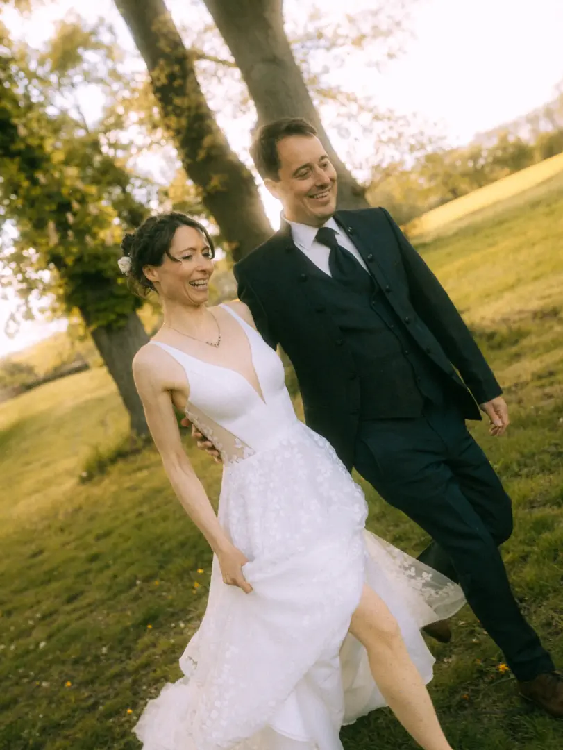 Ein glückliches Brautpaar spaziert lachend durch eine sonnige Wiese. Die Braut trägt ein elegantes, weißes Spitzenkleid mit Beinschlitz, der Bräutigam einen dunklen Anzug. Eine authentische Aufnahme von einem Lovestory Fotograf in Schwerin und MV.