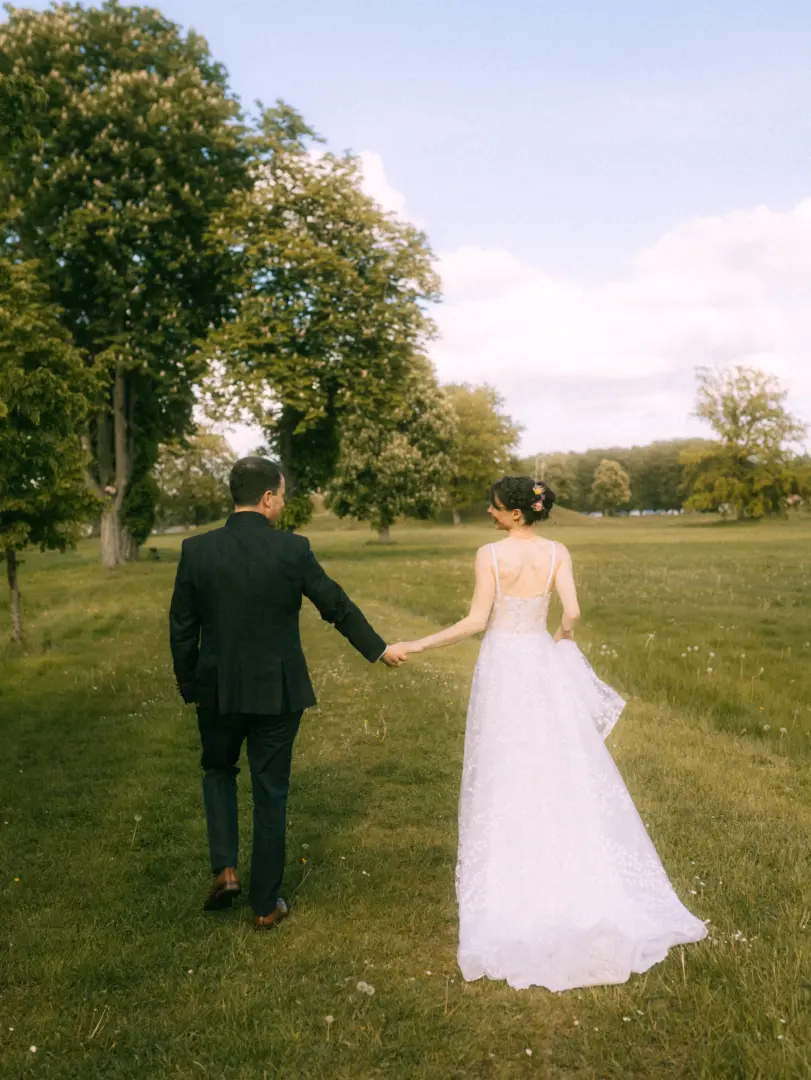 Ein Brautpaar geht Hand in Hand über eine grüne Wiese vor einer malerischen Baumkulisse. Die Braut trägt ein elegantes weißes Hochzeitskleid und blickt den Bräutigam im dunklen Anzug lächelnd an. Eine romantische Aufnahme vom Hochzeitsfotograf in Schwerin und MV.