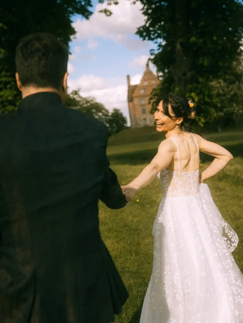 Eine strahlende Braut in einem weißen Spitzenkleid zieht ihren Bräutigam im schwarzen Anzug lachend über eine sonnige Wiese vor einem historischen Schloss. Ein romantischer Moment, eingefangen von einem Hochzeitsfotograf in Schwerin und MV.