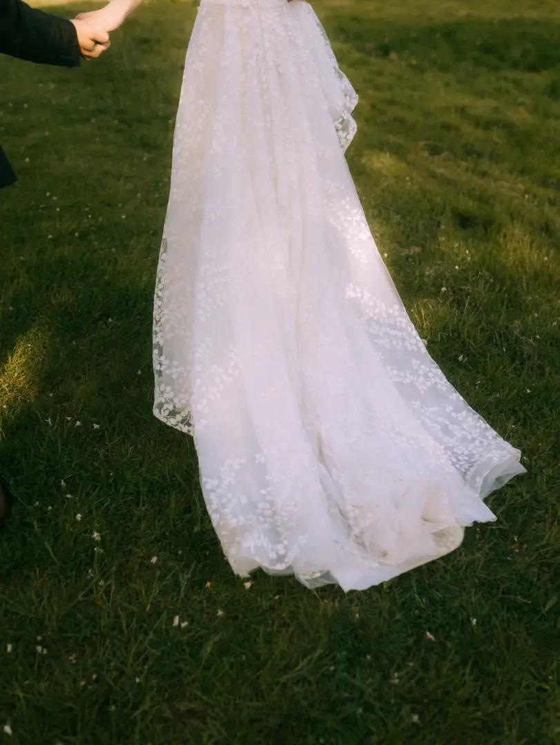 Eine Braut in einem eleganten, mit zarten floralen Mustern bestickten Brautkleid hält händchenhaltend die Hand ihres Partners, während sie gemeinsam über eine grüne Wiese spazieren. Stimmungsvolle Aufnahme von einem Hochzeitsfotograf in Schwerin und MV.