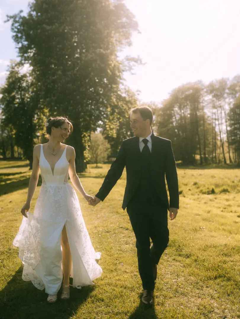 Ein glückliches Brautpaar hält händchenhaltend inne und blickt sich bei einem romantischen Spaziergang auf einer sonnendurchfluteten Wiese an. Die Braut trägt ein elegantes weißes Hochzeitskleid, der Bräutigam einen klassischen Anzug. Ein stimmungsvoller Moment vom Hochzeitsfotograf in Schwerin und MV.