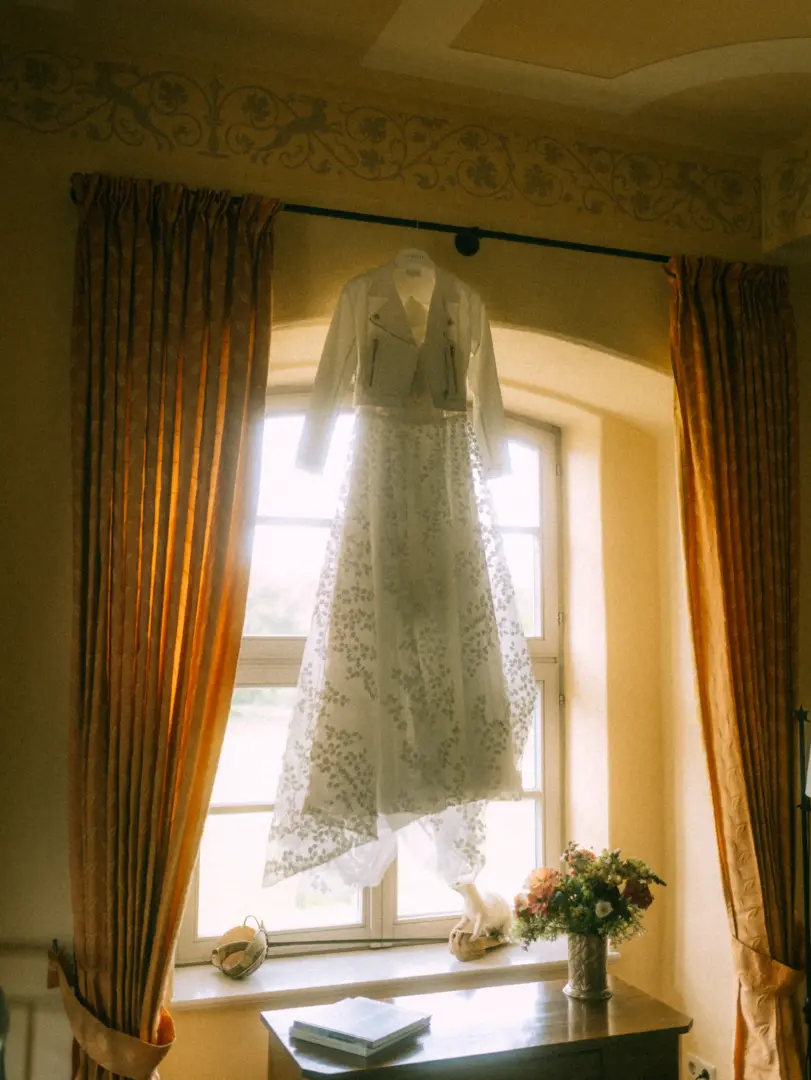 Ein weißes Brautkleid mit passender Lederjacke hängt elegant vor einem hellen Fenster, eingerahmt von goldgelben Vorhängen. Auf dem Fensterbrett steht ein Blumenstrauß. Eine stimmungsvolle Aufnahme von einem Hochzeitsfotograf in Schwerin und MV.
