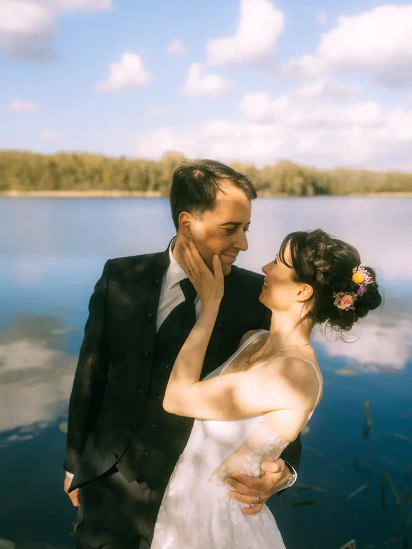 Ein frisch vermähltes Paar in inniger Umarmung an einem ruhigen See. Die Braut trägt ein weißes Spitzenkleid mit Blumen im Haar und berührt zärtlich das Gesicht des Bräutigams im dunklen Anzug. Eine romantische Szene, eingefangen von einem Hochzeitsfotograf in Schwerin und MV.