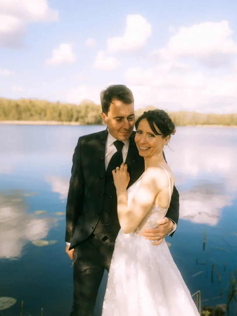 Ein glückliches Brautpaar posiert für ein romantisches Foto an einem ruhigen See unter blauem Himmel. Die Braut trägt ein weißes Spitzenkleid, während der Bräutigam einen dunklen Anzug trägt. Eine wunderschöne Aufnahme vom Lovestory Fotograf in Schwerin und MV.