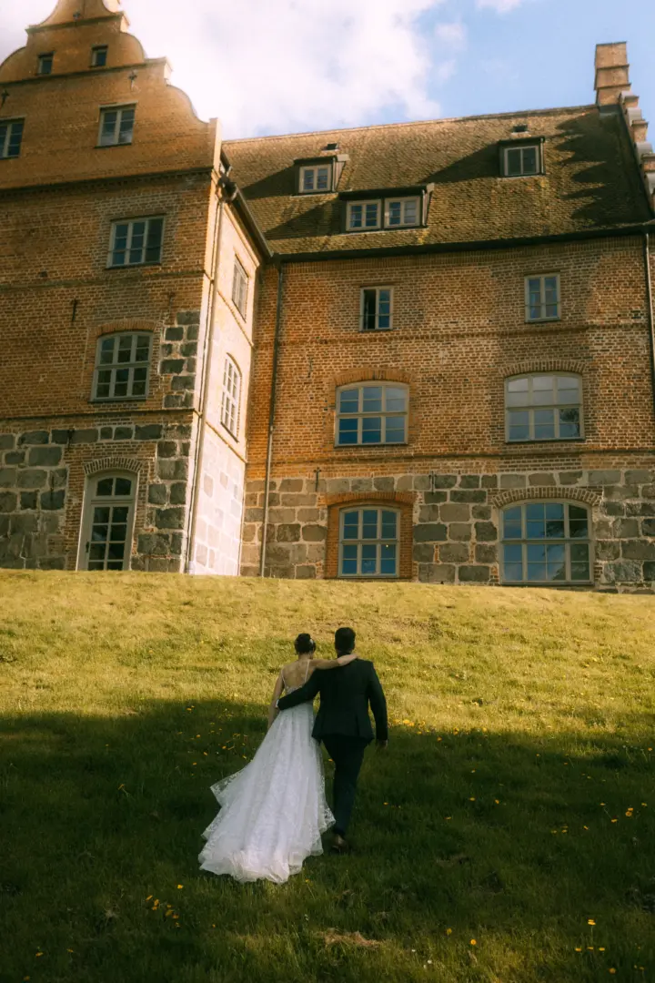 Ein Brautpaar geht Hand in Hand einen grünen Hügel vor einem historischen Backsteinschloss hinauf. Die Braut trägt ein elegantes weißes Hochzeitskleid, der Bräutigam einen dunklen Anzug. Eine romantische Szene, eingefangen von einem Hochzeitsfotograf in Schwerin und MV.