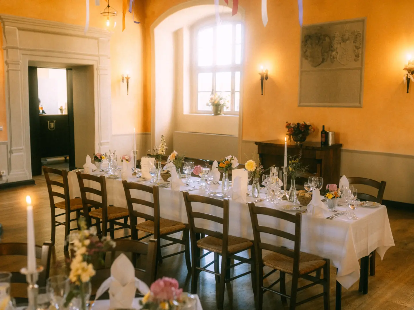 Festlich gedeckte lange Tafel in einem historischen Saal mit warmem, goldenem Licht, dekoriert mit Blumenarrangements, Kerzen und weißen Servietten für eine elegante Feier, fotografiert von einem Hochzeitsfotograf in Schwerin und MV.