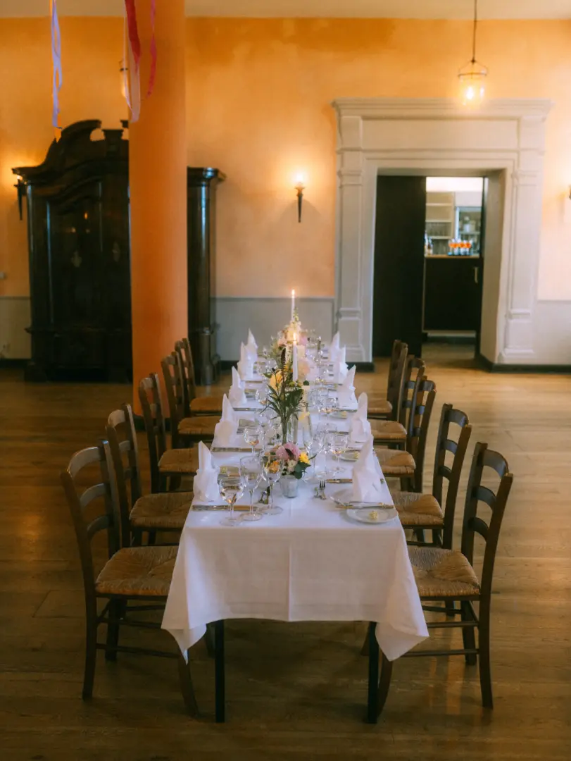 Festlich gedeckte, lange Tafel in einem historischen Saal mit warmen orangefarbenen Wänden und Holzboden, perfekt für eine Hochzeit. Die Tischdekoration umfasst weiße Servietten, Blumenarrangements und Kerzen. Hochzeitsfotograf in Schwerin und MV.