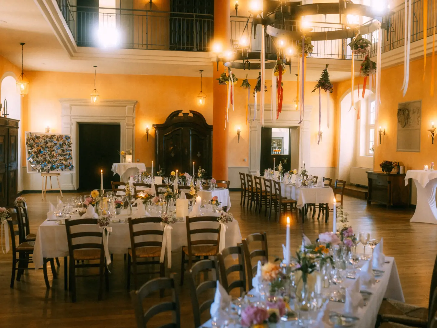 Festlich gedeckte Hochzeitstafeln in einem historischen Saal mit hohen, ockergelben Wänden, eleganten Kronleuchtern und hängenden Blumendekorationen. Ein stimmungsvolles Ambiente für eine Feier, festgehalten von einem Hochzeitsfotograf in Schwerin und MV.