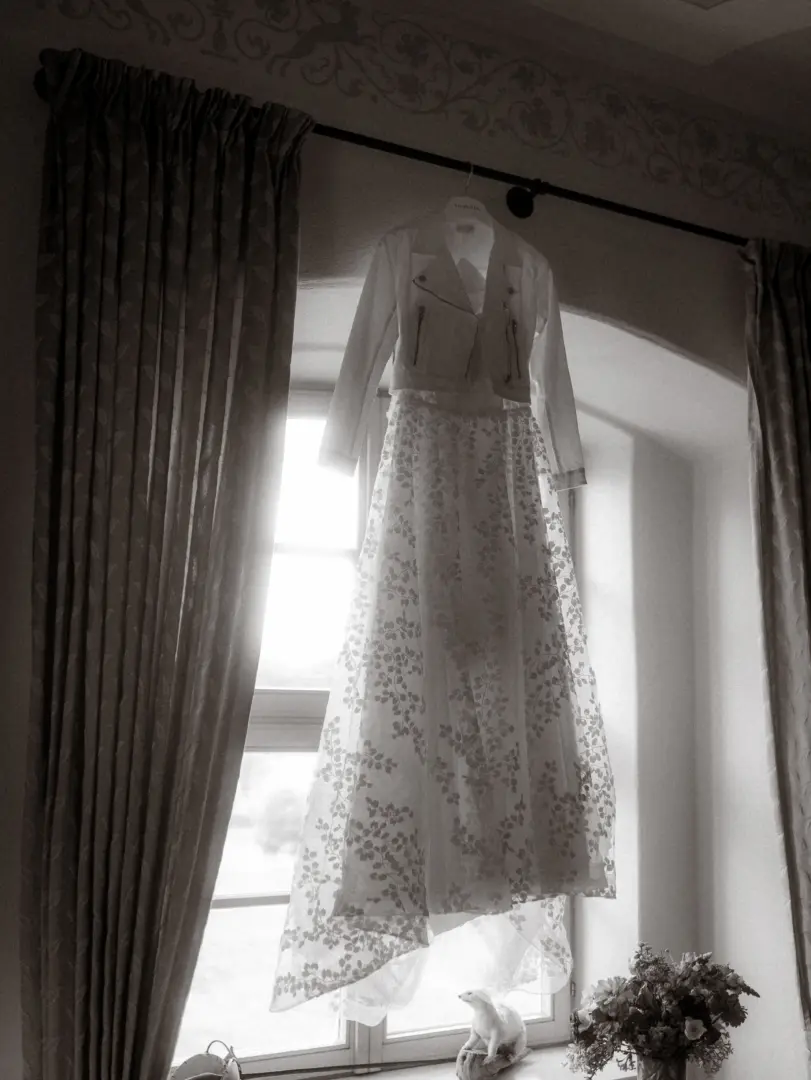 Ein elegantes Brautkleid mit floralem Spitzenmuster und eine dazu passende weiße Bikerjacke hängen vor einem hellen Fenster. Das stimmungsvolle Hochzeitsfoto wurde vom Hochzeitsfotograf in Schwerin und MV aufgenommen und fängt die Vorfreude auf den großen Tag ein.
