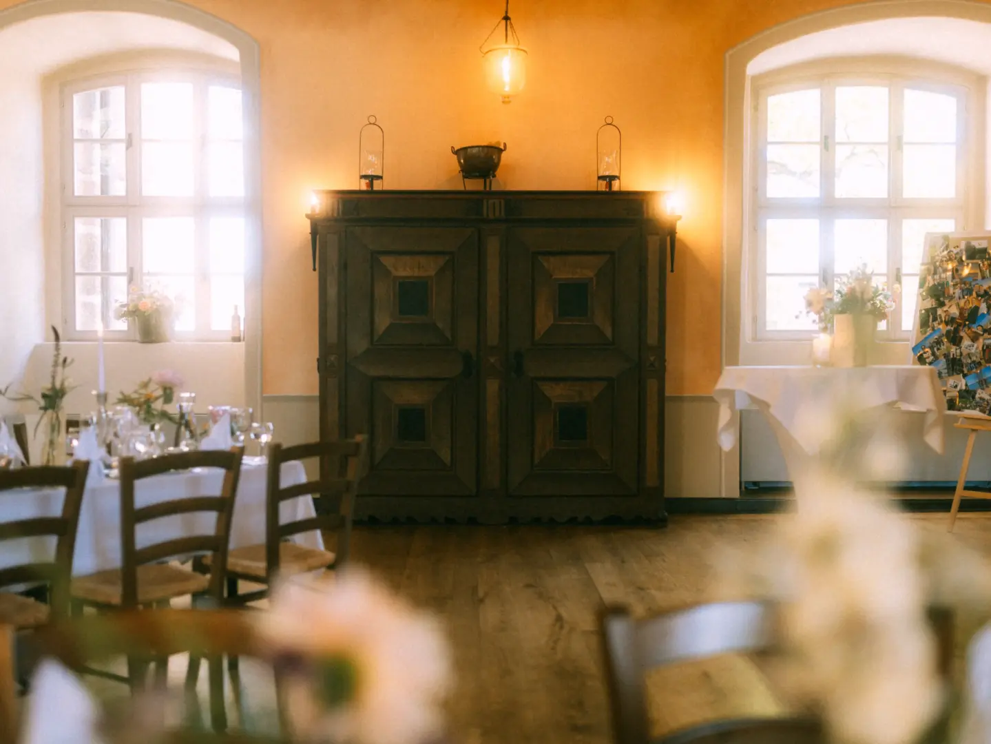 Ein festlich gedeckter Raum für eine Hochzeitsfeier mit einem großen antiken Holzschrank in der Mitte, flankiert von zwei hellen Fenstern und warmem Kerzenlicht. Die Atmosphäre ist einladend und elegant, festgehalten von einem Hochzeitsfotograf in Schwerin und MV.