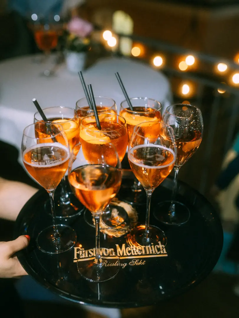 Eine Hand hält ein schwarzes Serviertablett mit dem Logo „Fürst von Metternich Riesling Sekt“, auf dem mehrere gefüllte Sekt- und Aperol-Spritz-Gläser mit Strohhalmen bei einer Abendveranstaltung stehen. Stimmungsvolle Lichterketten im Hintergrund sorgen für ein elegantes Ambiente.