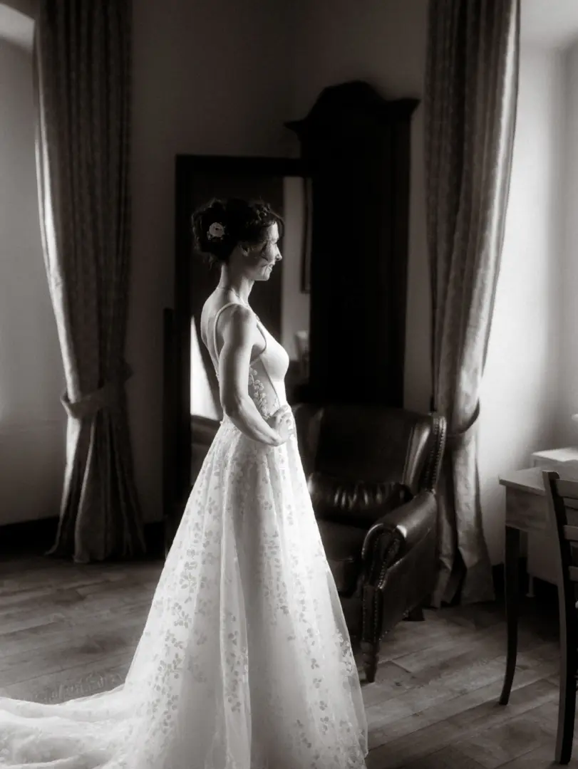 Eine Braut im eleganten, floral bestickten Hochzeitskleid steht in einem stilvollen Raum vor einem Fenster, während sie ihren Hochzeitstag genießt. Die Schwarz-Weiß-Aufnahme fängt diesen intimen Moment perfekt ein – ein Beispiel für die Arbeit als Hochzeitsfotograf in Schwerin und MV.