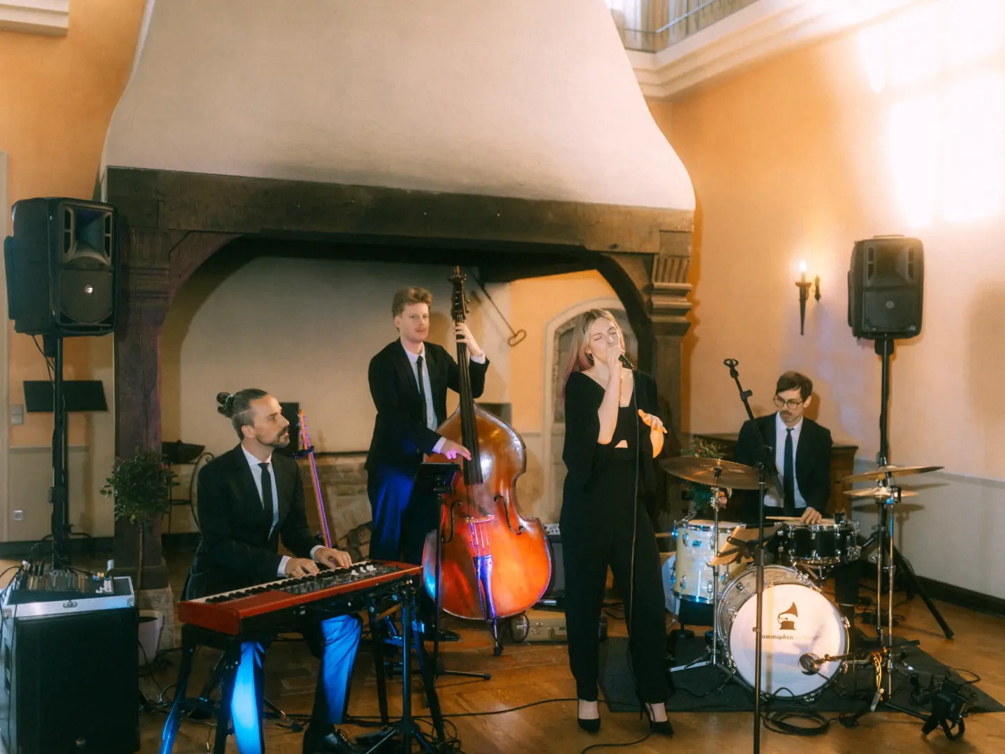 Eine vierköpfige Jazzband bei einem Auftritt auf einer Hochzeit: Ein Pianist, ein Kontrabassist, eine Sängerin und ein Schlagzeuger in eleganten Anzügen und schwarzer Kleidung spielen vor einem historischen Kamin. Hochzeitsfotograf in Schwerin und MV.
