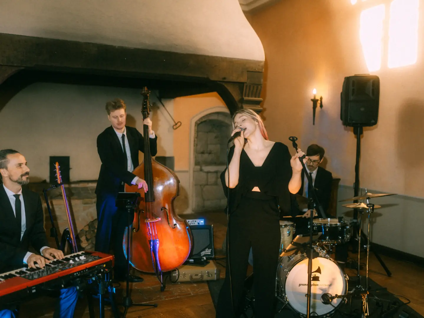 Eine Jazzband performt live bei einer Hochzeitsfeier in einem historischen Raum mit Kamin. Die Sängerin im schwarzen Outfit steht im Mittelpunkt, begleitet von Musikern an Kontrabass, Keyboard und Schlagzeug. Hochzeitsfotograf in Schwerin und MV.