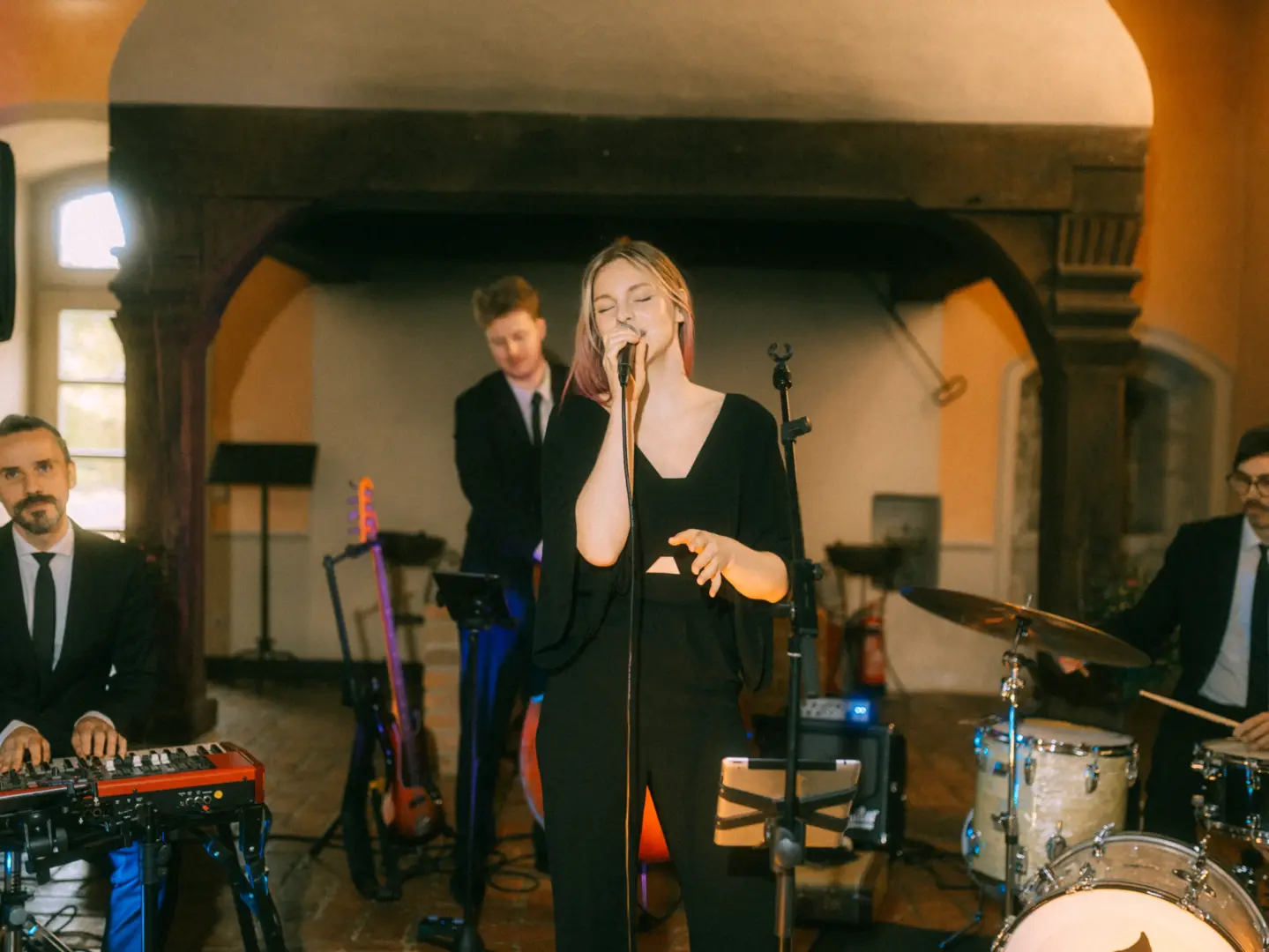 Eine Live-Band tritt bei einer Hochzeit auf: Eine Sängerin mit rosa Strähnen singt mit geschlossenen Augen, begleitet von einem Keyboarder, einem Bassisten und einem Schlagzeuger in Anzügen. Hochzeitsfotograf in Schwerin und MV.