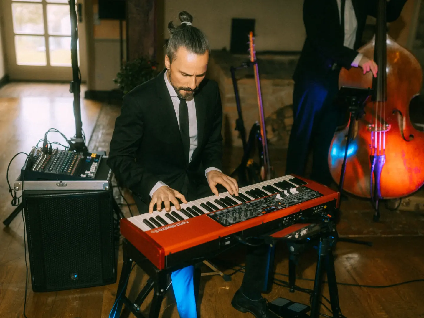 Ein Musiker mit Dutt spielt konzentriert auf einem roten Nord Electro 5 Keyboard während einer Veranstaltung. Im Hintergrund begleitet ihn ein Kontrabassist. Professionelle Live-Musik sorgt für eine elegante Atmosphäre, festgehalten von Ihrem Hochzeitsfotograf in Schwerin und MV.