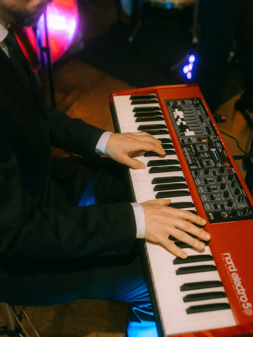 Ein Musiker in einem eleganten schwarzen Anzug spielt auf einem roten Nord Electro 5 Keyboard bei einer Hochzeitsfeier. Die stimmungsvolle Beleuchtung verleiht dem Moment eine festliche Atmosphäre. Hochzeitsfotograf in Schwerin und MV.