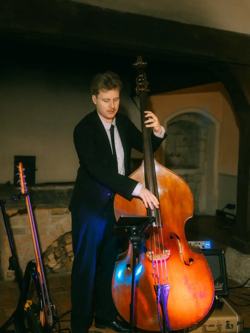Ein junger Musiker im eleganten schwarzen Anzug spielt konzentriert einen Kontrabass bei einer Hochzeitsfeier. Im Hintergrund ist ein rustikaler Kamin zu sehen, während das Lichtspiel für eine stimmungsvolle Atmosphäre sorgt – festgehalten von Ihrem Hochzeitsfotograf in Schwerin und MV.