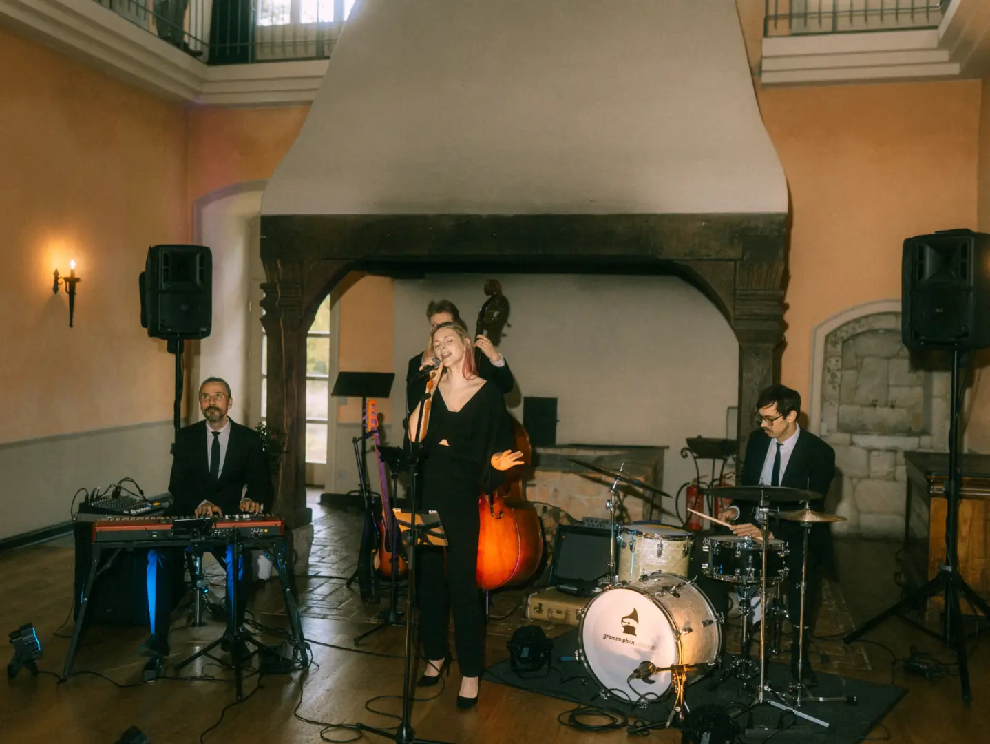 Eine Jazzband tritt bei einer Hochzeit auf: Eine Sängerin in schwarzem Outfit steht im Mittelpunkt, begleitet von einem Keyboarder, einem Kontrabassisten und einem Schlagzeuger in einem eleganten Saal. Hochzeitsfotograf in Schwerin und MV.