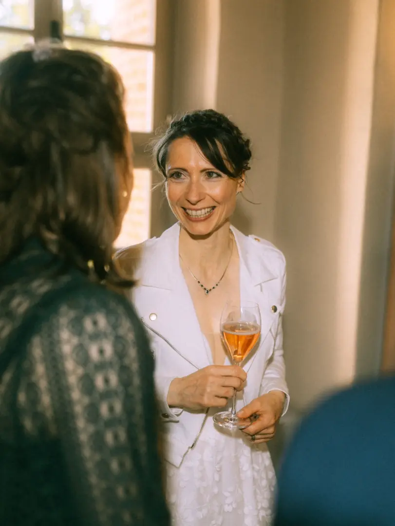 Eine strahlende Braut in einer weißen Lederjacke hält ein Glas Rosé-Sekt in der Hand und unterhält sich herzlich bei einer Hochzeitsfeier. Ein Hochzeitsfotograf in Schwerin und MV fängt diesen authentischen Moment der Freude und Geselligkeit in einer hellen, festlichen Atmosphäre ein.