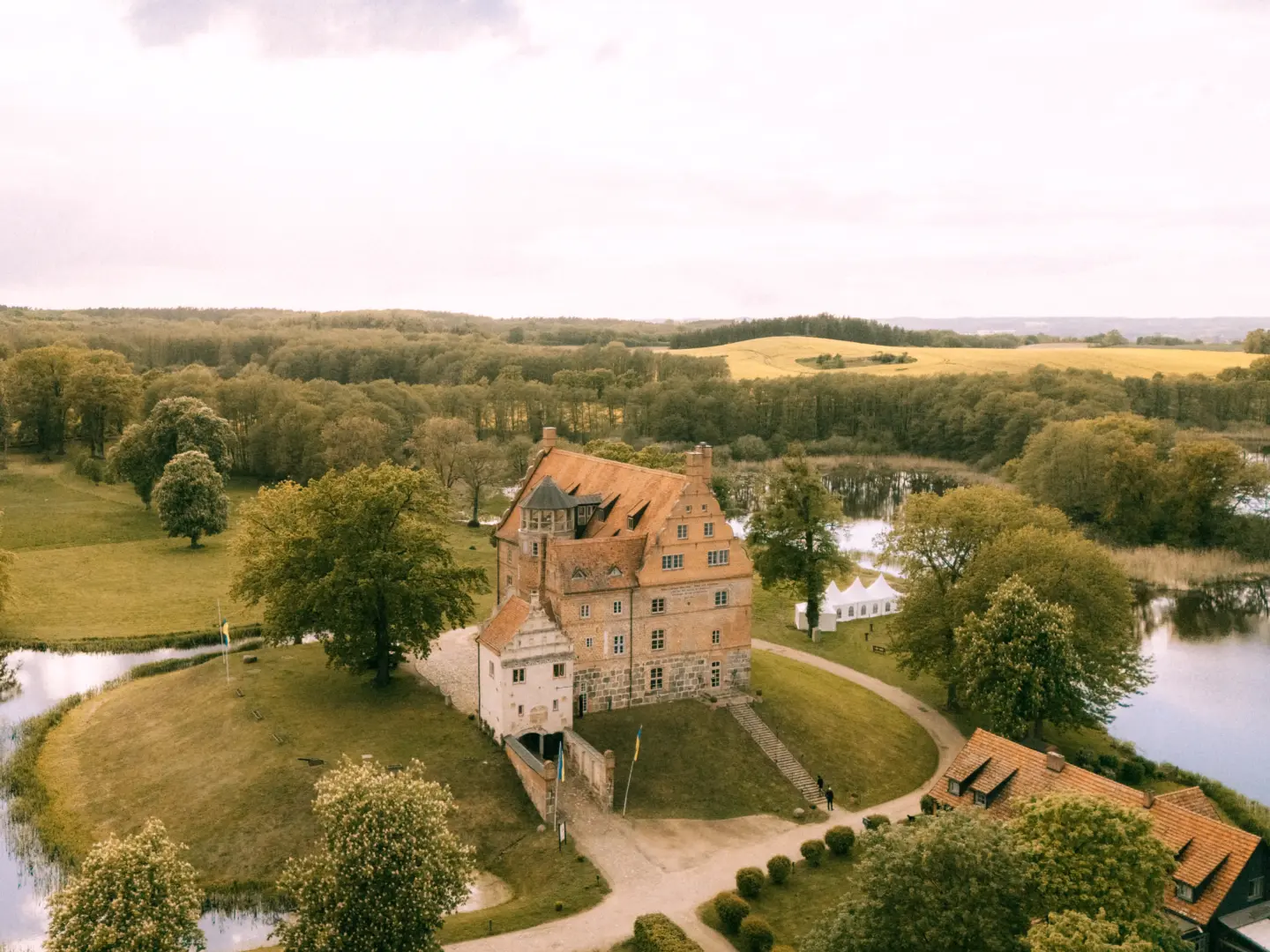 Luftaufnahme von Schloss Ulrichshusen, einem historischen Backsteinschloss, das malerisch auf einer Halbinsel inmitten einer Seenlandschaft liegt. Ein weißes Festzelt im Hintergrund deutet auf eine exklusive Hochzeitslocation hin, fotografiert von einem Hochzeitsfotograf in Schwerin und MV.