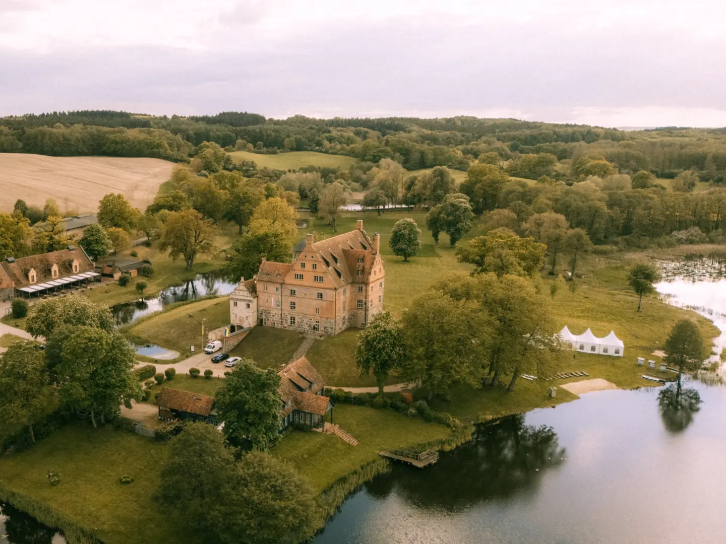 Luftaufnahme eines historischen Backsteinschlosses an einem See in einer idyllischen Parklandschaft, perfekt geeignet als Kulisse für den Hochzeitsfotograf in Schwerin und MV. Das Schloss mit seinen Nebengebäuden und weißen Festzelten liegt eingebettet in eine grüne Naturkulisse.