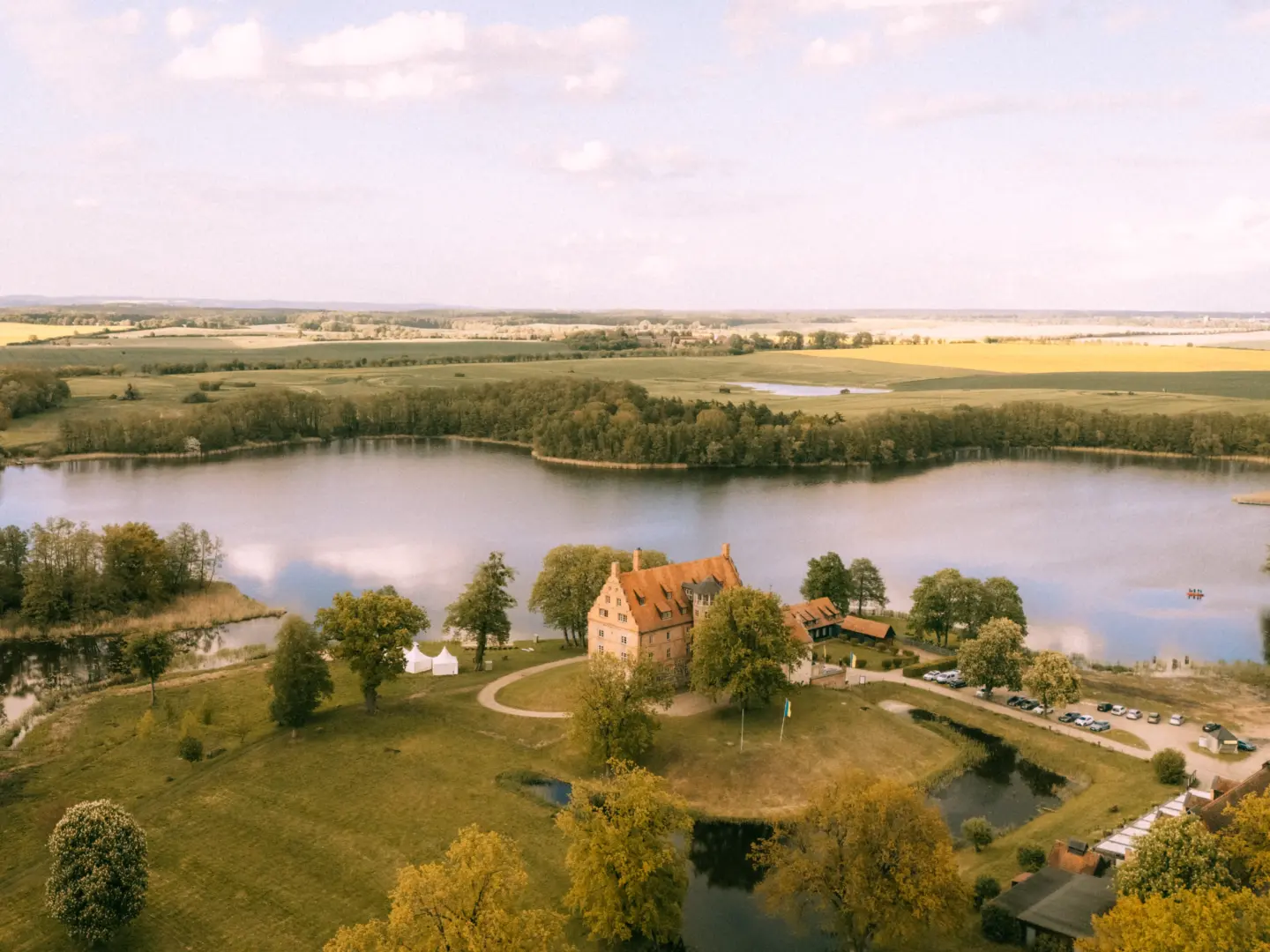 Luftaufnahme eines historischen Herrenhauses an einem ruhigen See in Mecklenburg-Vorpommern, umgeben von grünen Wiesen und Wäldern. Die idyllische Kulisse eignet sich perfekt für einen Hochzeitsfotograf Müritz und MV.