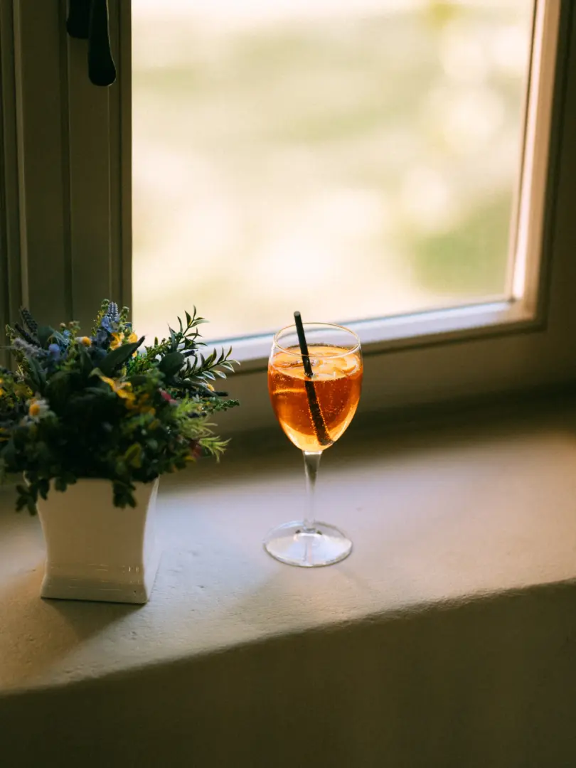 Ein erfrischender Aperol Spritz mit einer Orangenscheibe und einem schwarzen Strohhalm steht in einem eleganten Weinglas auf einer Fensterbank neben einem kleinen, bunten Blumenarrangement. Das warme Licht fällt sanft durch das Fenster und erzeugt eine entspannte, sommerliche Atmosphäre.