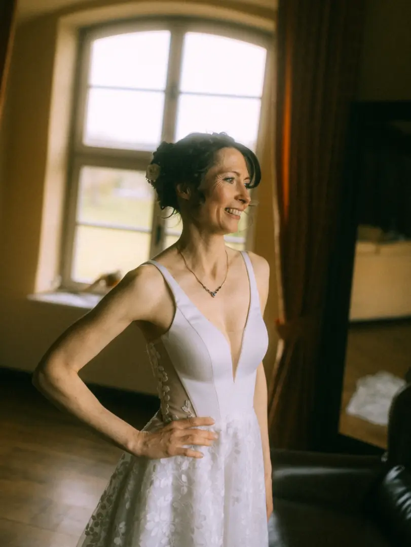 Eine strahlende Braut in einem eleganten weißen Brautkleid mit tiefem Ausschnitt und zarten Spitzenapplikationen steht lächelnd vor einem hellen Fenster. Das stimmungsvolle Porträt wurde von einem Hochzeitsfotograf in Schwerin und MV aufgenommen.