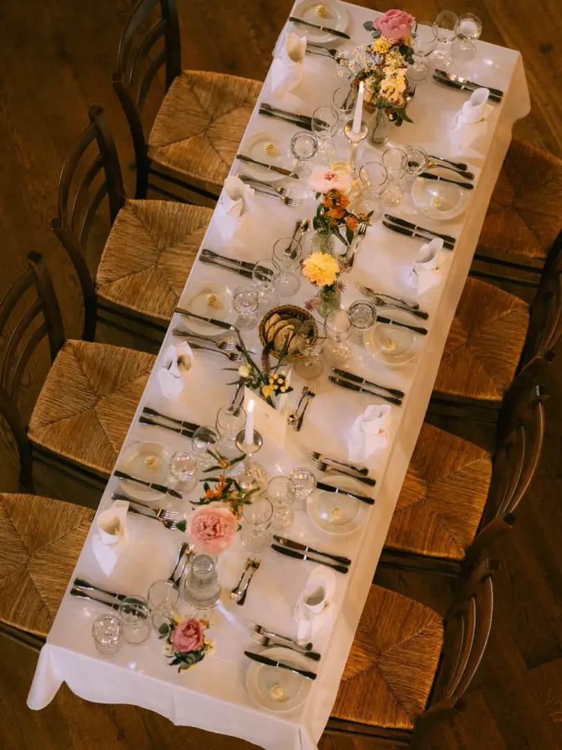 Festlich gedeckte lange Tafel für eine Hochzeitsfeier mit weißer Tischdecke, rustikalen Holzstühlen, eleganten Blumenarrangements in Pastelltönen und Kerzen. Professionelle Aufnahme durch einen Hochzeitsfotograf in Schwerin und MV, die das stilvolle Ambiente der Tischdekoration einfängt.