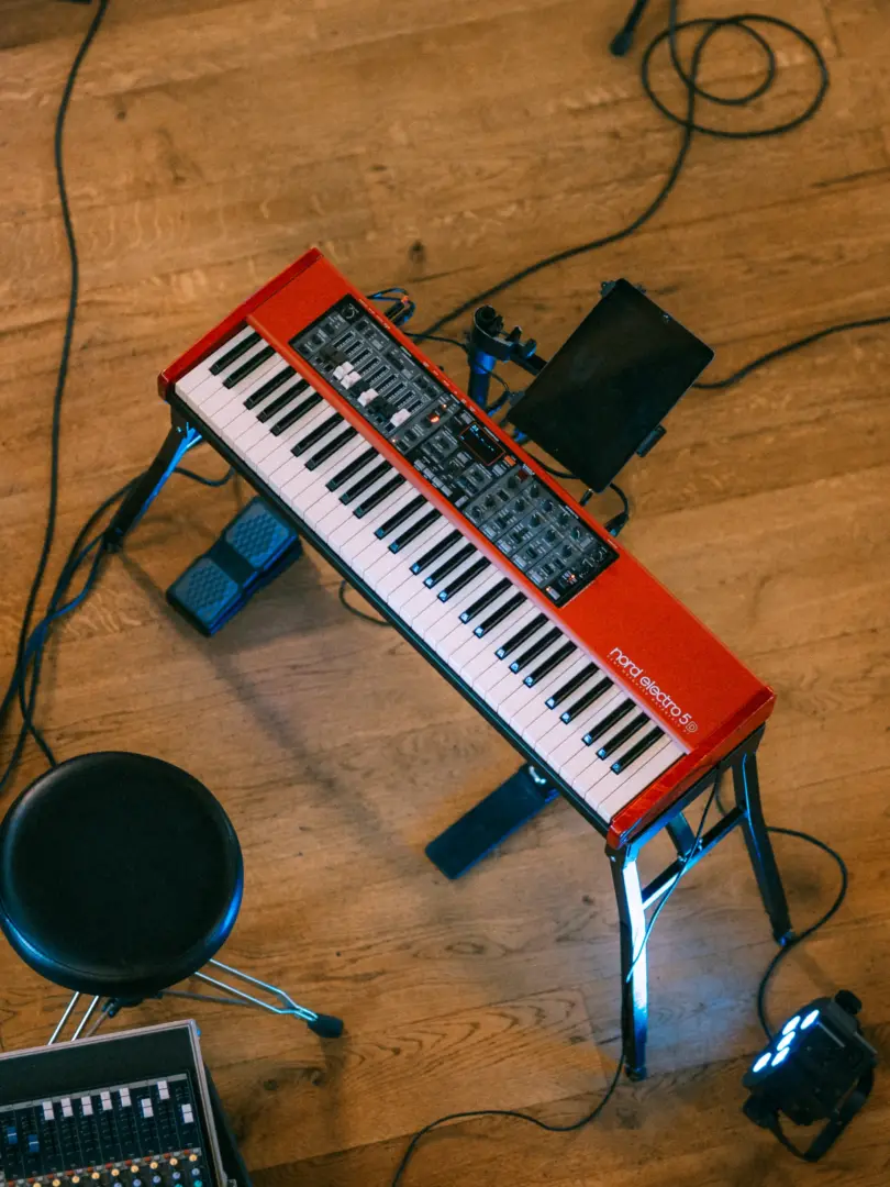 Ein rotes Nord Electro 5D Keyboard steht auf einem Ständer in einem Raum mit Holzboden. Neben dem Instrument befinden sich ein schwarzer Hocker, ein Fußpedal, ein Tablet auf einer Halterung und ein Mischpult, während ein kleiner Bühnenscheinwerfer den Boden dezent beleuchtet.