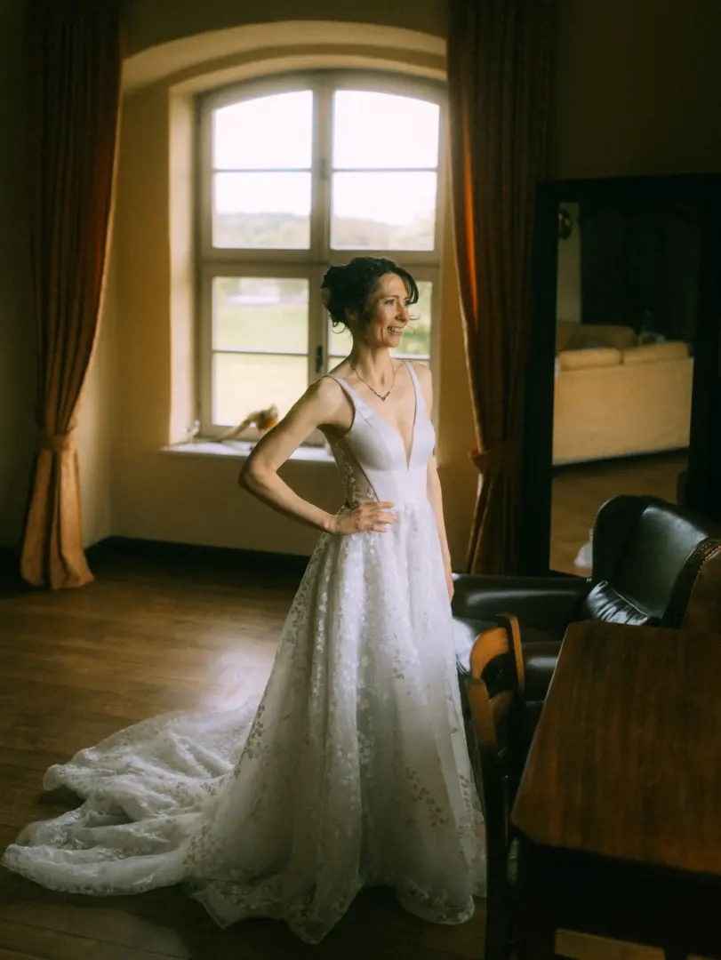 Eine strahlende Braut in einem eleganten, bodenlangen Hochzeitskleid mit floraler Spitze steht lächelnd vor einem großen Fenster in einem stilvollen Raum. Die Lichtstimmung unterstreicht die romantische Atmosphäre, eingefangen von einem Hochzeitsfotograf in Schwerin und MV.