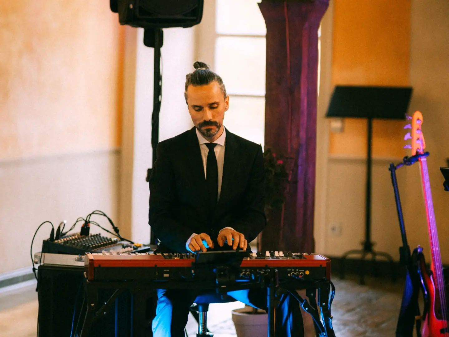 Ein konzentrierter Musiker mit Dutt spielt bei einer Hochzeitsfeier auf einem roten Stage-Piano. Er trägt einen eleganten schwarzen Anzug mit Krawatte, während im Hintergrund ein Bass auf einem Ständer steht. Professionelle Begleitung durch einen Hochzeitsfotograf in Schwerin und MV.