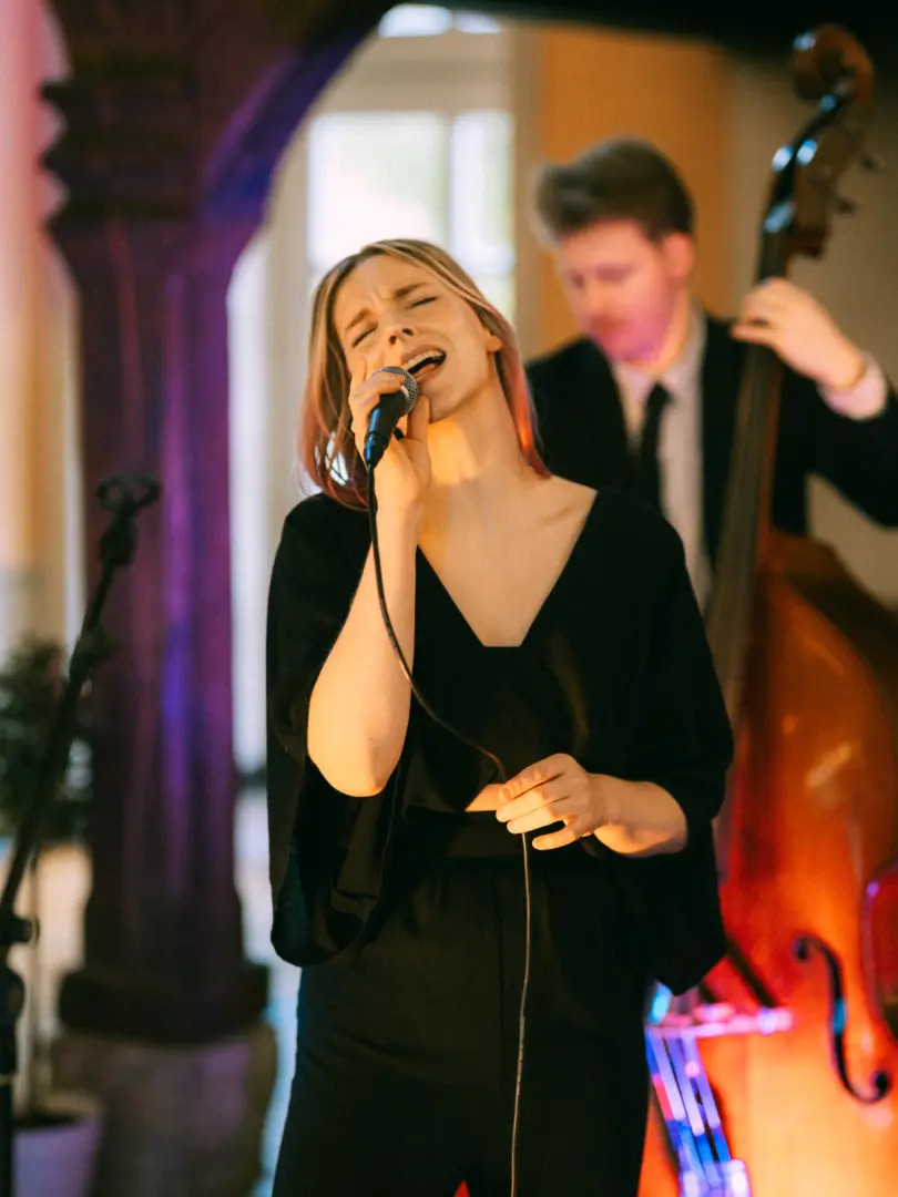 Eine leidenschaftlich singende Frau mit Mikrofon in einem schwarzen Outfit, begleitet von einem Kontrabassisten im Hintergrund bei einer festlichen Veranstaltung. Professionelle Aufnahme von einem Hochzeitsfotograf in Schwerin und MV.