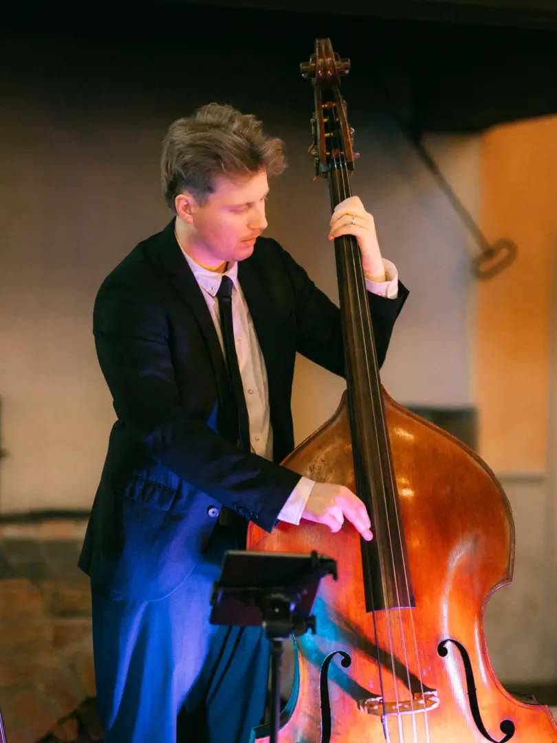 Ein Kontrabassspieler in einem eleganten schwarzen Anzug konzentriert sich während eines Auftritts auf sein Instrument. Das stimmungsvolle Licht in Blau- und Rottönen unterstreicht die musikalische Atmosphäre. Hochzeitsfotograf in Schwerin und MV.