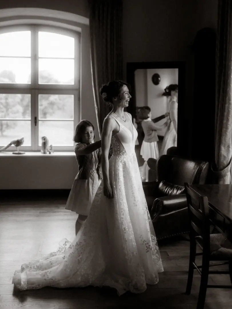 Eine strahlende Braut in einem eleganten, bestickten Hochzeitskleid wird von einem kleinen Mädchen beim Ankleiden unterstützt. Die Szene fängt einen emotionalen Moment in einem hellen Raum ein, während sich die Braut im Spiegel betrachtet. Hochzeitsfotograf in Schwerin und MV.