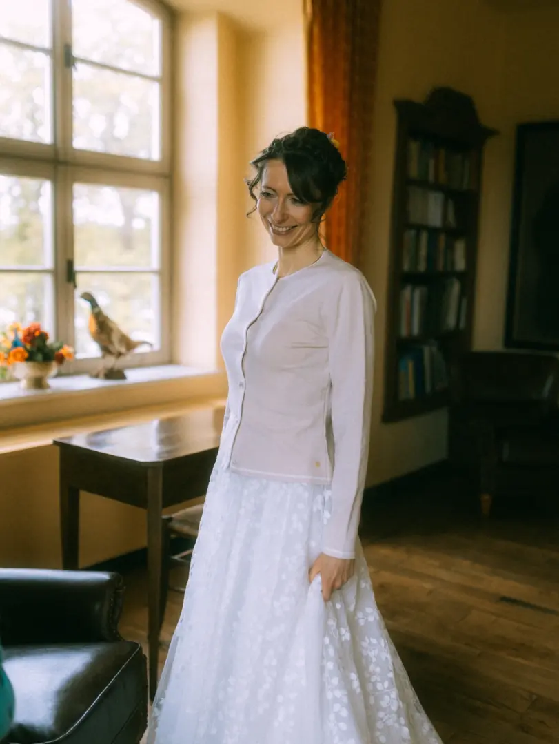Eine lächelnde Braut in einem weißen Spitzenrock und einer hellen Strickjacke steht in einem gemütlichen, lichtdurchfluteten Zimmer mit einem Bücherregal im Hintergrund. Dieses stimmungsvolle Bild wurde von einem Hochzeitsfotograf in Schwerin und MV aufgenommen.