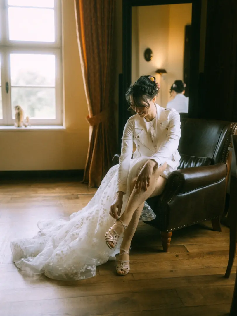 Eine Braut in einem weißen Spitzenkleid und einer modernen Bikerjacke sitzt in einem Ledersessel und zieht ihre glitzernden Sandalen an. Die Szene wirkt elegant und entspannt, festgehalten von einem Hochzeitsfotograf in Schwerin und MV.