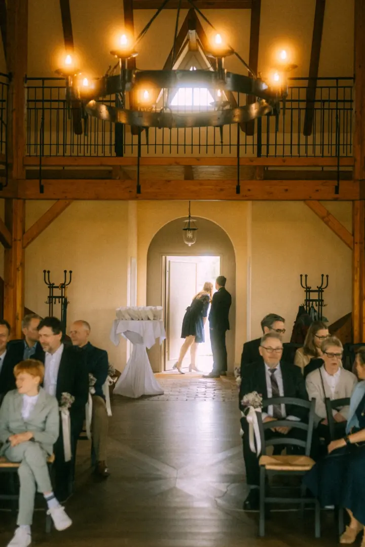 Ein Brautpaar steht in der hellen Türöffnung einer rustikalen Hochzeitslocation, während die Hochzeitsgäste im Vordergrund gespannt auf die Zeremonie warten. Stimmungsvolle Aufnahme von einem Hochzeitsfotograf in Schwerin und MV.