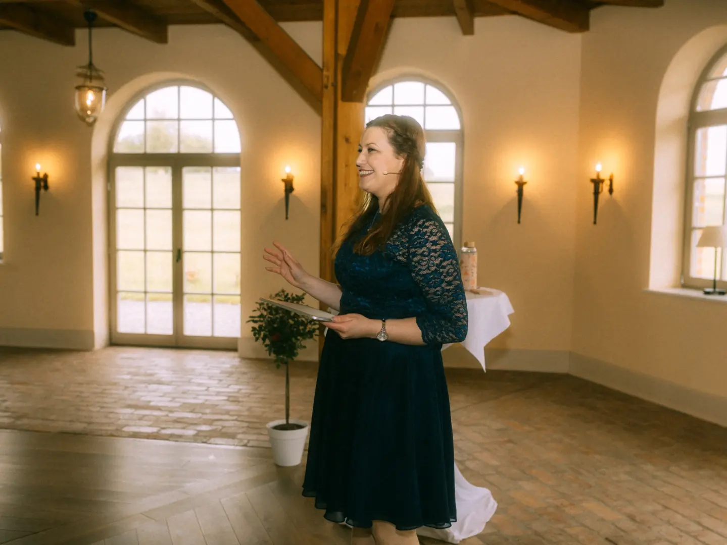 Eine lächelnde Rednerin in einem eleganten dunkelblauen Spitzenkleid hält eine Rede in einem hellen, rustikalen Saal mit großen Rundbogenfenstern. Der Raum bietet eine stimmungsvolle Kulisse für Hochzeitsfotograf in Schwerin und MV.