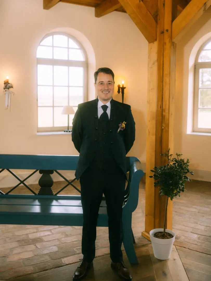 Ein Bräutigam im eleganten schwarzen Anzug mit Weste und Ansteckblume steht lächelnd vor einer blauen Holzbank in einem hellen, rustikalen Raum mit großen Rundbogenfenstern. Hochzeitsfotograf in Schwerin und MV.