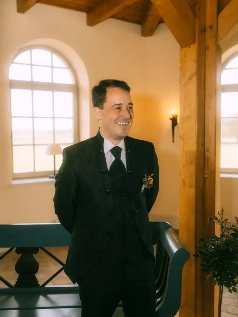 Ein glücklicher Bräutigam in einem eleganten schwarzen Anzug mit Weste und Ansteckblume lächelt während seiner Trauung. Im Hintergrund sorgt ein helles Bogenfenster für warmes Licht in der rustikalen Hochzeitslocation. Hochzeitsfotograf in Schwerin und MV.