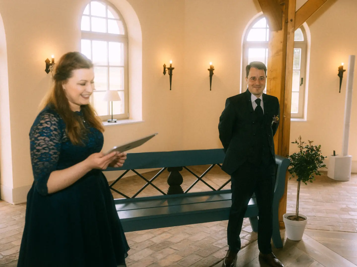 Eine Frau in einem dunkelblauen Spitzenkleid hält ein Tablet in den Händen und lächelt, während sie vor einem Mann im dunklen Anzug steht. Die Szene findet in einem hellen, festlichen Raum statt. Hochzeitsfotograf in Schwerin und MV.