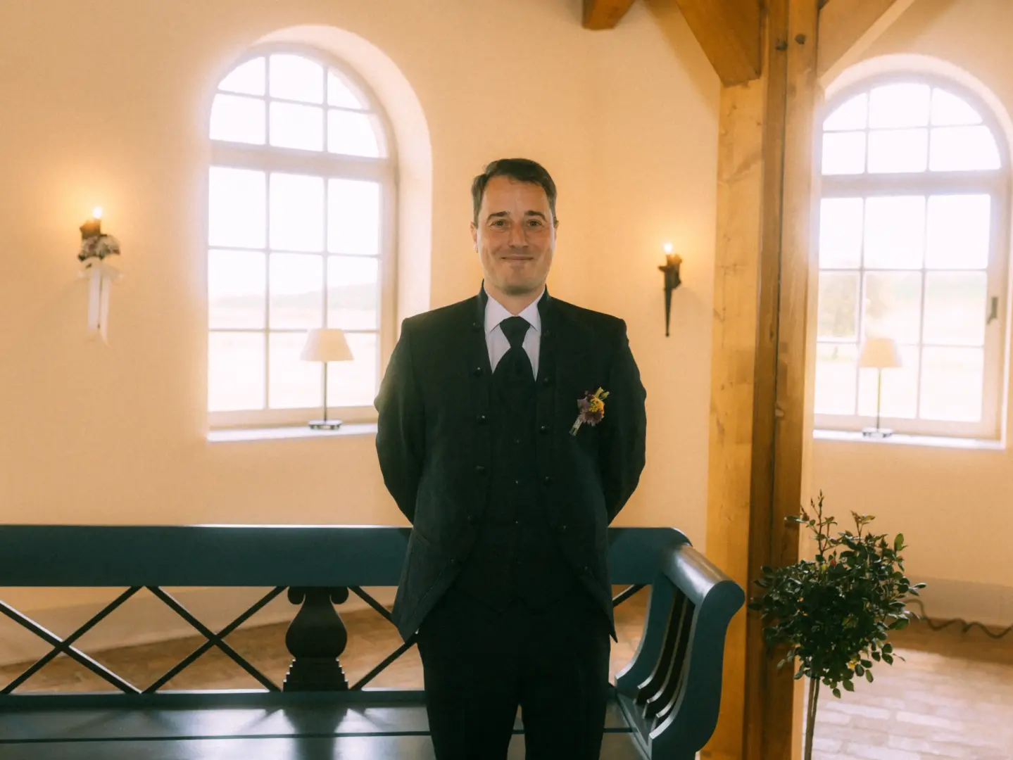 Ein Bräutigam im eleganten schwarzen Anzug mit Weste und Ansteckblume steht lächelnd in einer hellen, rustikalen Hochzeitslocation vor zwei großen Rundbogenfenstern. Hochzeitsfotograf in Schwerin und MV.
