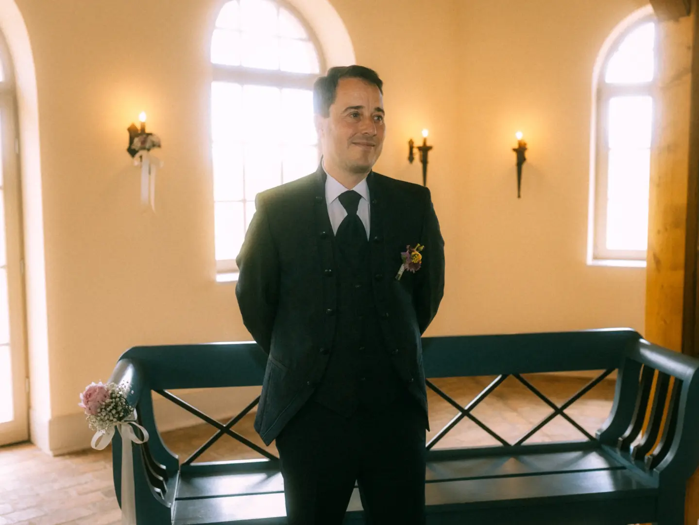Ein Bräutigam wartet in einem hellen, festlich geschmückten Raum auf seine Braut. Er trägt einen eleganten schwarzen Anzug mit Weste und Blumenanstecker. Das Ambiente ist warm und einladend, aufgenommen von einem Hochzeitsfotograf in Schwerin und MV.