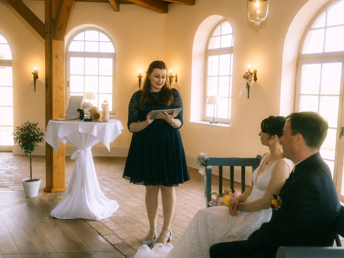 Eine Traurednerin in einem dunkelblauen Kleid liest während einer Zeremonie von einem Tablet vor, während ein Brautpaar ihr aufmerksam zuhört. Die Szene findet in einem hellen, eleganten Raum mit Rundbogenfenstern statt, aufgenommen von einem Hochzeitsfotograf in Schwerin und MV.