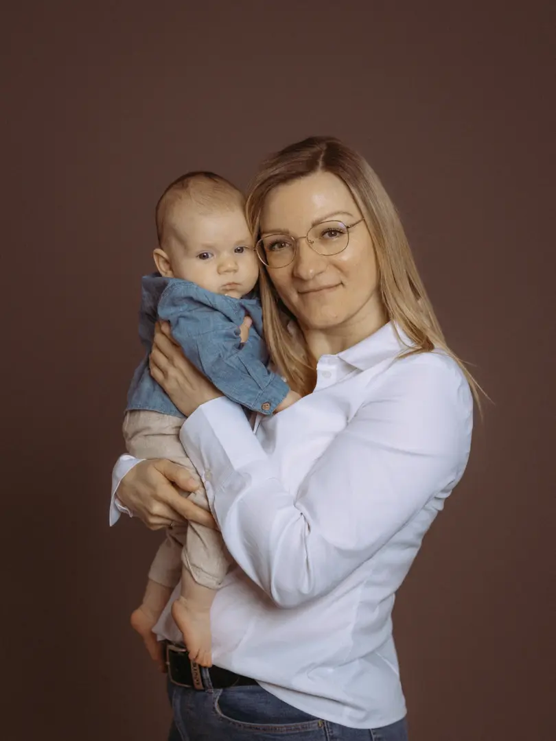Eine blonde Frau mit Brille hält liebevoll ihr Baby auf dem Arm in einem professionellen Fotostudio in Schwerin. Beide schauen direkt in die Kamera vor einem schlichten, braunen Hintergrund. Das Kind trägt ein blaues Hemd und helle Hosen, die Mutter ein weißes Hemd.