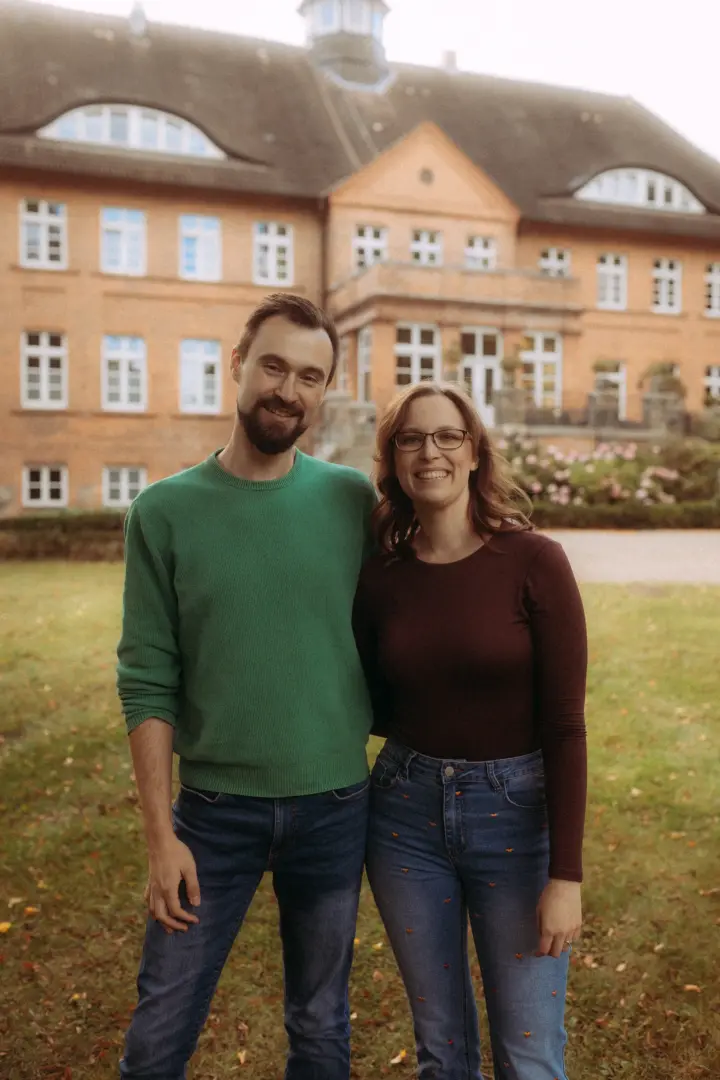 Ein glückliches Paar lächelt in einem Park vor einem historischen Backsteingebäude. Der Mann trägt einen grünen Pullover, die Frau ein weinrotes Oberteil. Diese authentischen Pärchenfotos in Schwerin fangen eine entspannte Lovestory ein.