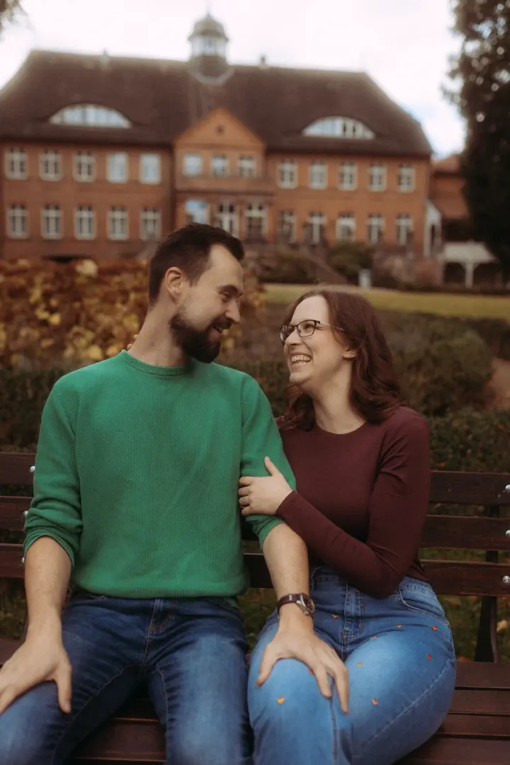 Ein glückliches Paar sitzt eng beieinander auf einer Parkbank vor einem historischen Backsteingebäude. Der Mann trägt einen grünen Pullover, die Frau ein bordeauxrotes Oberteil. Sie lachen sich liebevoll an, während sie den Moment genießen. Pärchenfotos Schwerin.