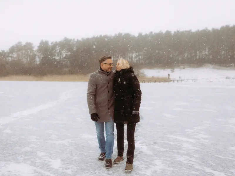 Ein glückliches Paar spaziert eng umschlungen über einen zugefrorenen, schneebedeckten See, während es sanft schneit. Die beiden genießen einen romantischen Wintertag in der Natur. Authentische Pärchenfotos Schwerin für unvergessliche Erinnerungen.